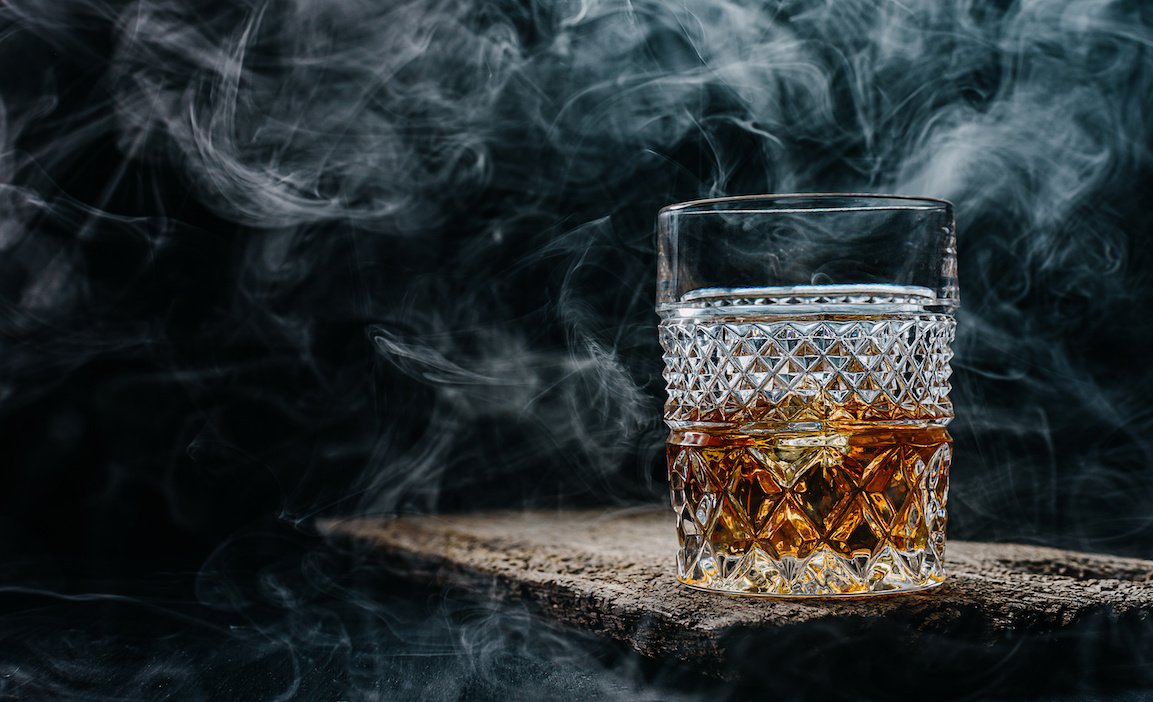 glass of whiskey with ice on a wooden table surrounded by smoke